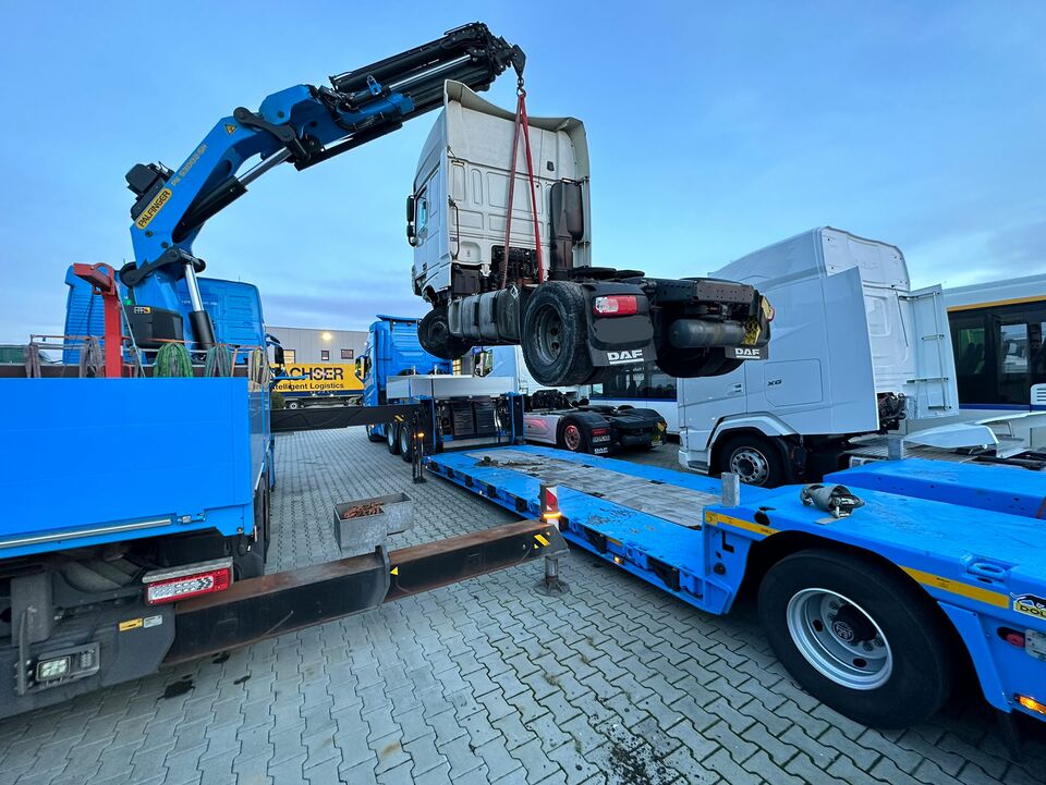 Ein blauer Kran hebt das Führerhaus eines LKW auf einen Tieflader in einem Industriegelände, umgeben von weiteren LKWs und Anhängern.