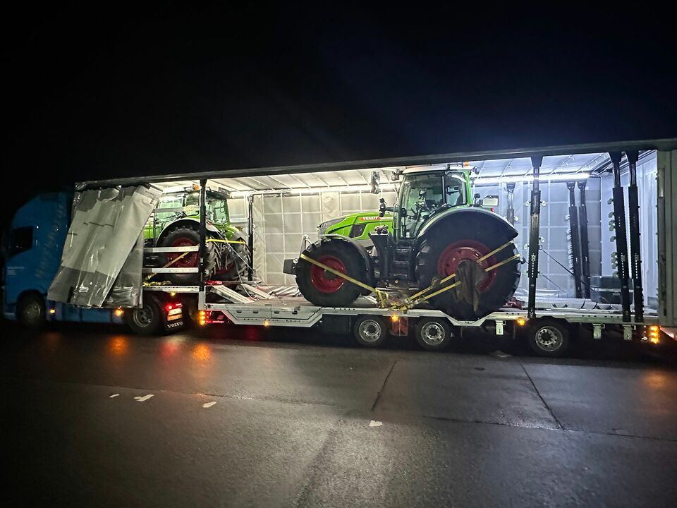 Ein blauer LKW transportiert zwei grüne Traktoren in einem beleuchteten Anhänger während der Nacht auf einer Straße.