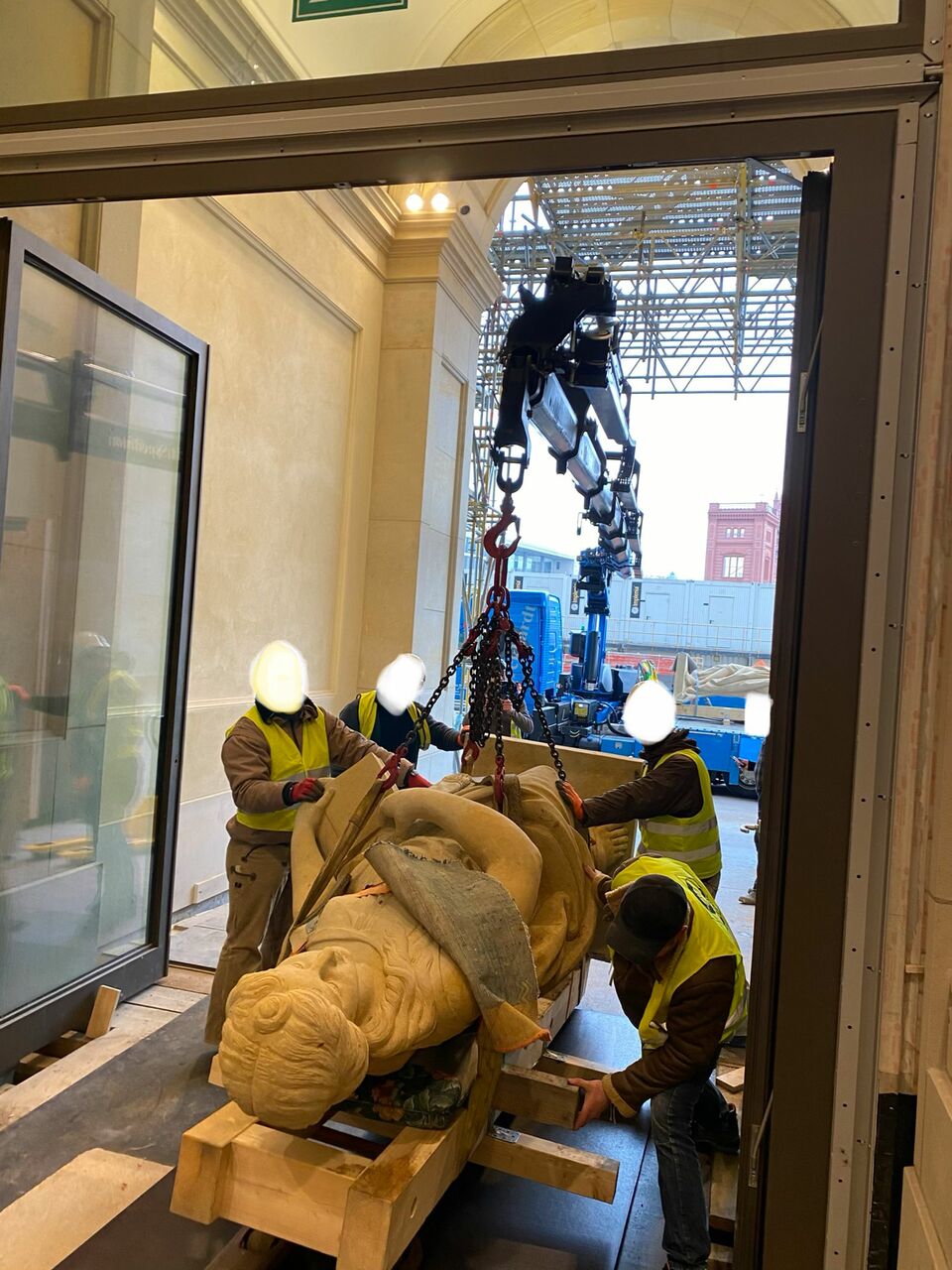 Arbeiter sichern eine große Skulptur, die mit einem Kran durch ein Gebäude transportiert wird, begleitet von präziser Kranführung und Hebetechnik.