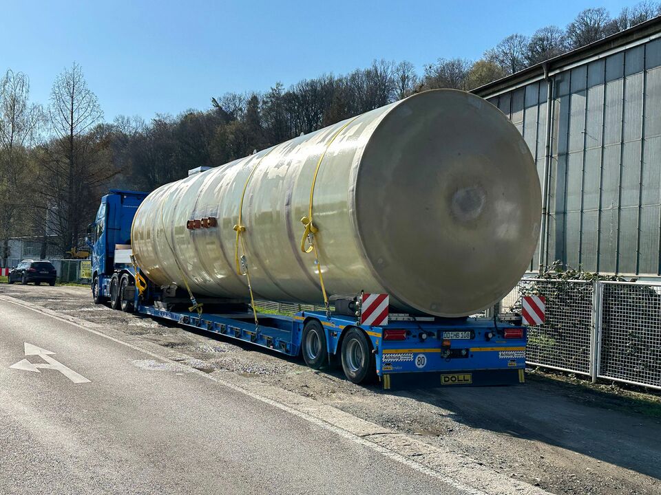 Ein großer zylinderförmiger Tank wird sicher auf einem blauen Tieflader-LKW transportiert, geparkt neben einer Industriehalle auf einer Landstraße.