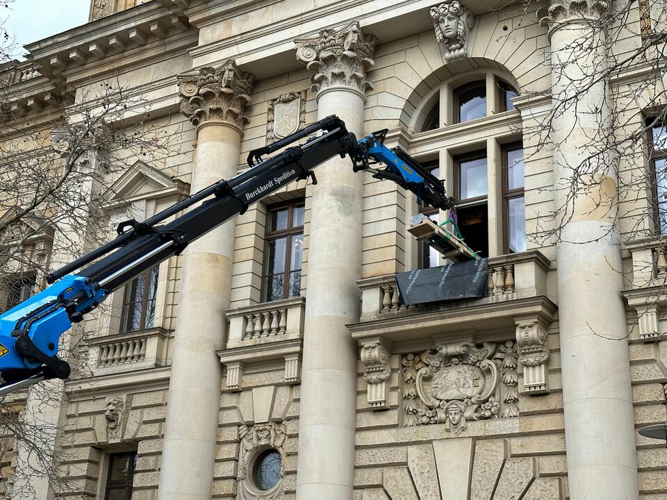 Ein blauer Kran hebt sorgfältig eine Ladung durch ein geöffnetes Fenster eines historischen Gebäudes mit dekorativen Säulen und Fassadenschmuck.