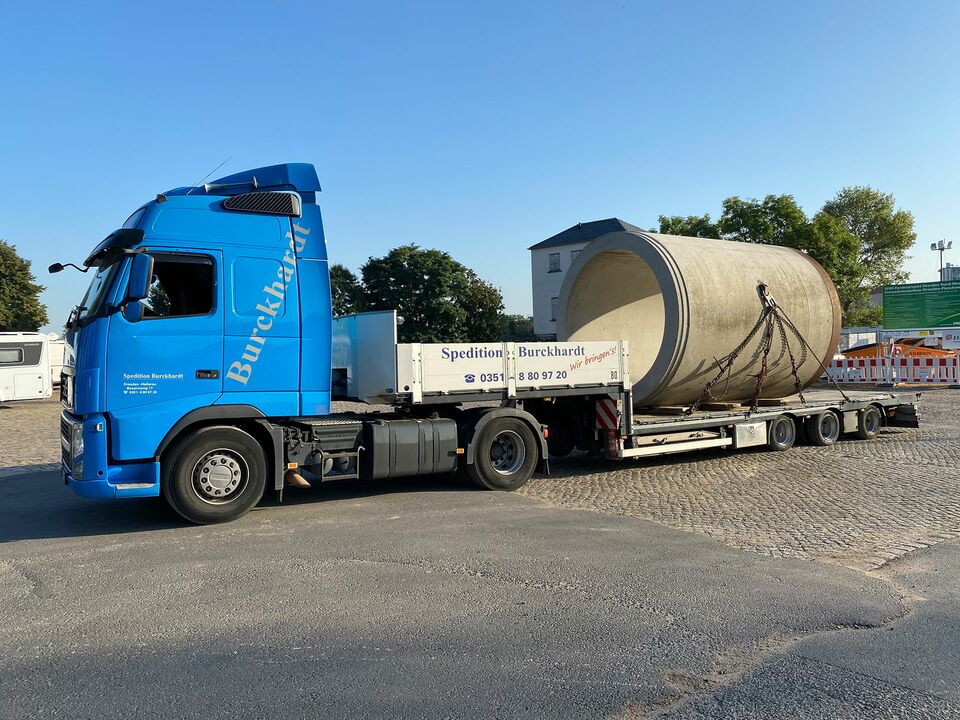 Ein blauer LKW transportiert ein massives, zylinderförmiges Betonrohr auf einem Tieflader über einen gepflasterten Platz.