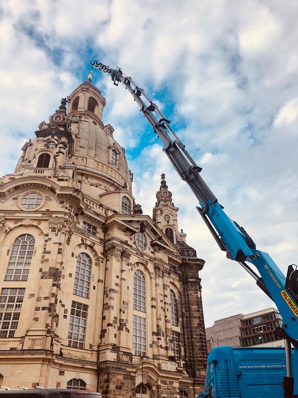 Ein blauer Kran arbeitet an der Spitze der historischen Frauenkirche in Dresden, vor einem leicht bewölkten Himmel.