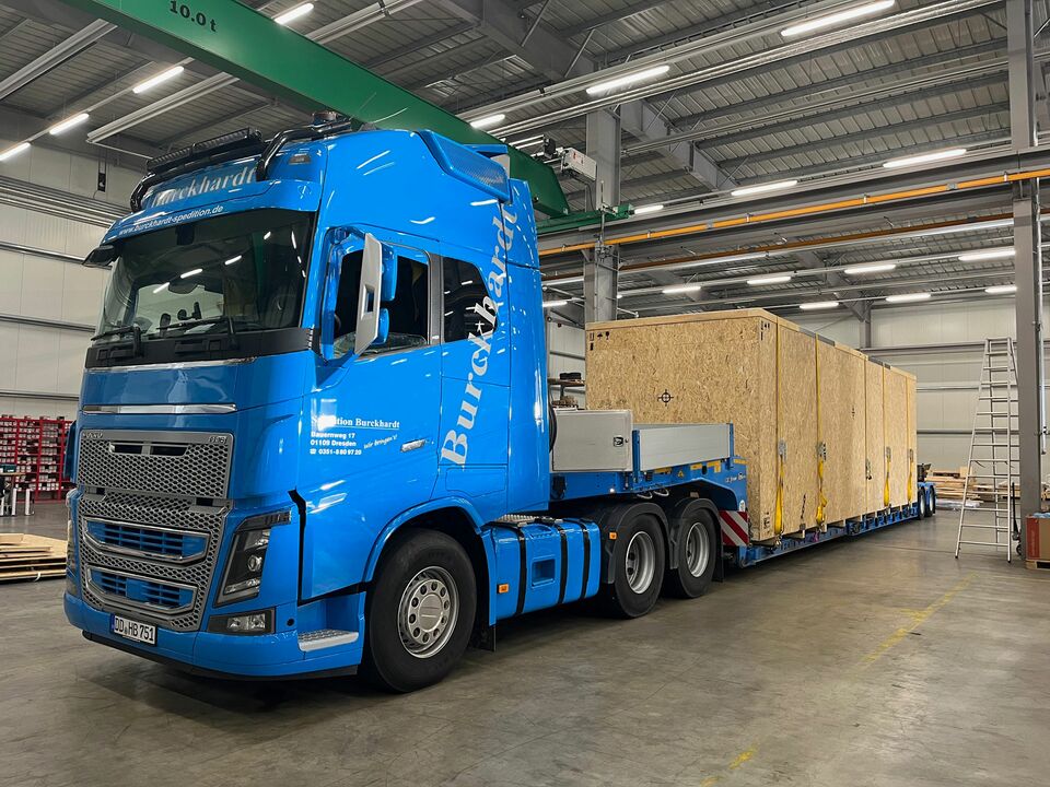 Ein blauer LKW transportiert massive Holzkisten auf einem Tieflader innerhalb einer Industriehalle mit Kränen und Beleuchtung.