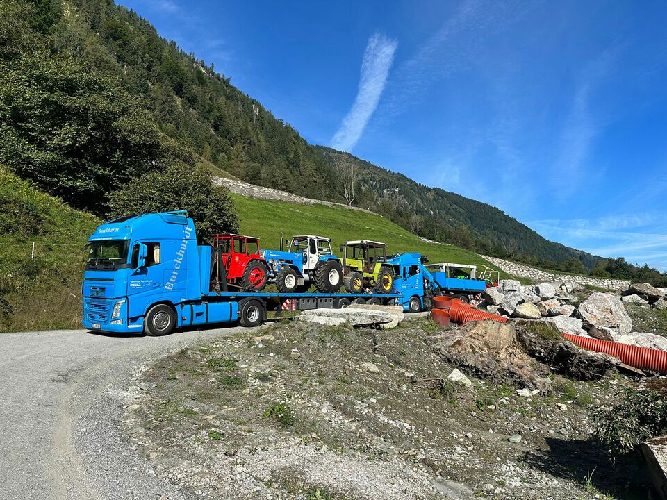 Ein blauer LKW transportiert mehrere Traktoren auf einem Tieflader durch eine alpine Landschaft mit grünen Hängen und blauem Himmel.