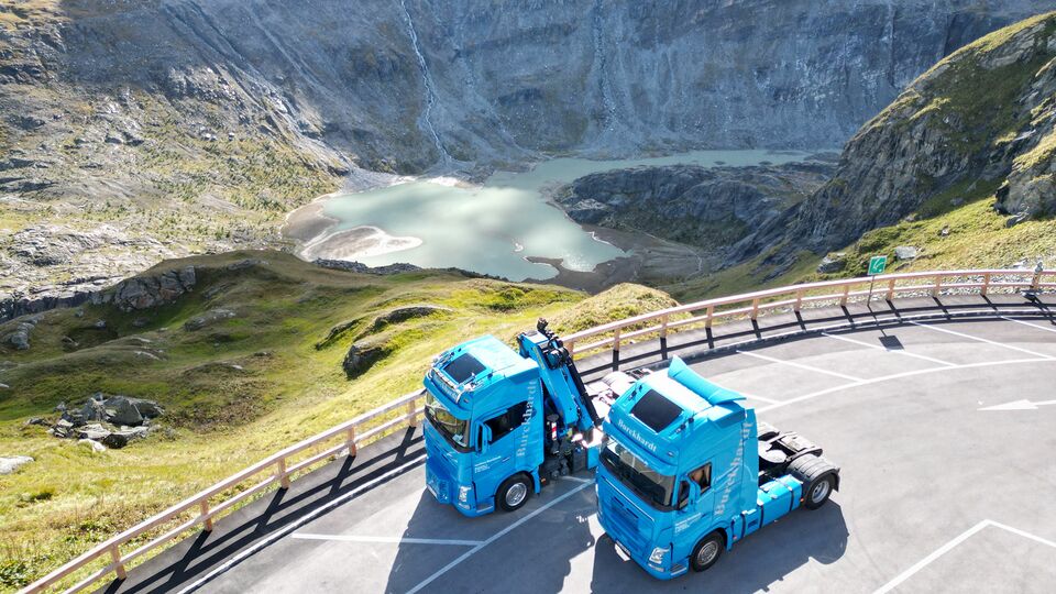 Zwei blaue LKWs stehen auf einem Parkplatz mit Blick auf einen Gebirgssee, umgeben von steilen Felswänden und grünen Wiesen.