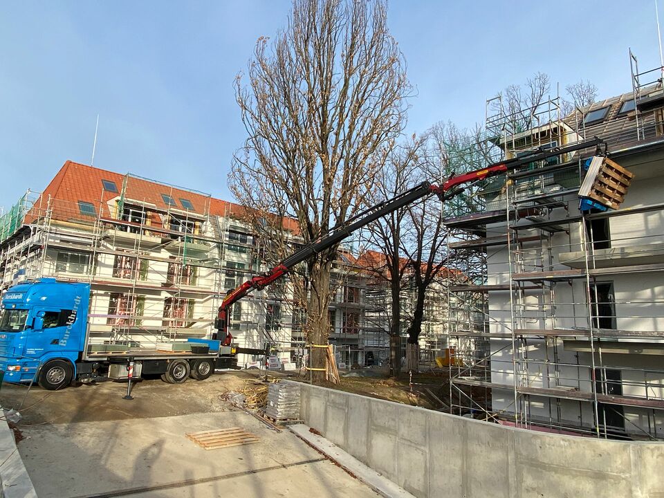 Ein blauer Kran hebt Baumaterialien auf das Dach eines mehrstöckigen Wohngebäudes, das mit Gerüsten umgeben ist, während im Hintergrund weitere Baustellen sichtbar sind.
