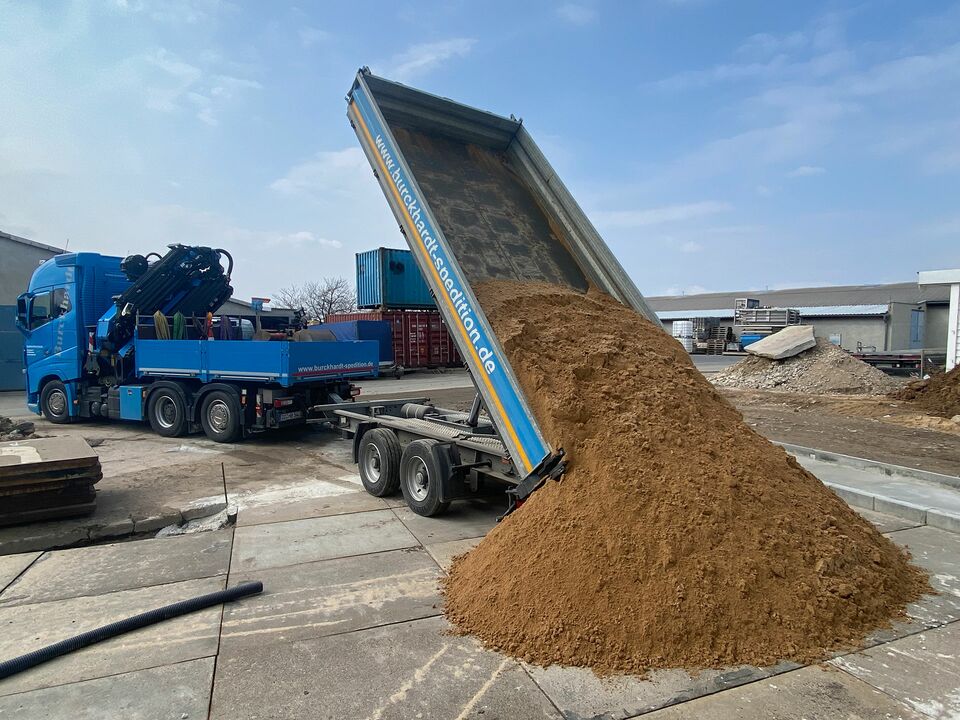 Ein blauer LKW kippt eine große Menge Sand von seinem Anhänger auf eine Baustelle, umgeben von Containern und Baumaterialien.