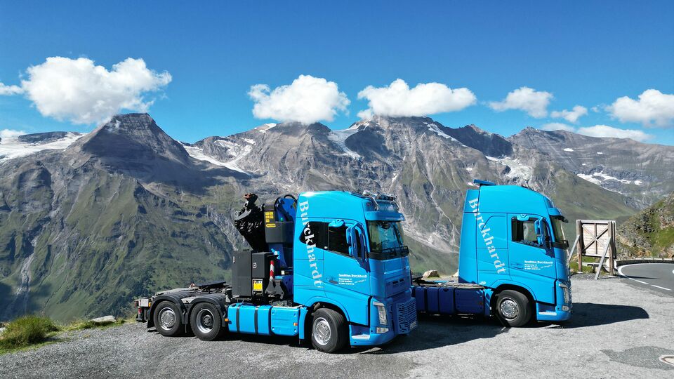 Zwei blaue LKWs parken vor der Kulisse majestätischer Alpenberge unter klarem, blauem Himmel.