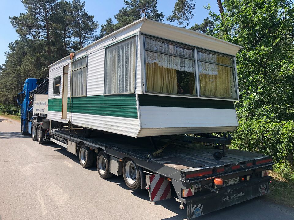 Ein Mobilheim mit grünen und weißen Seitenwänden wird auf einem Tieflader durch ein Waldgebiet transportiert.