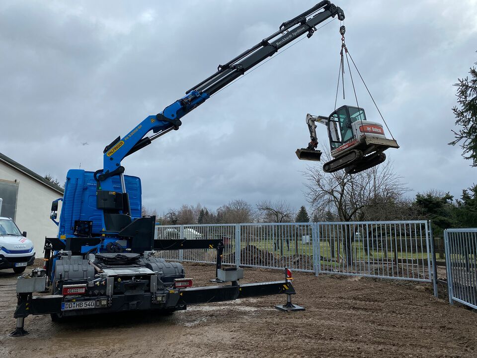 Ein Kran hebt einen kleinen Bagger auf einer Baustelle.