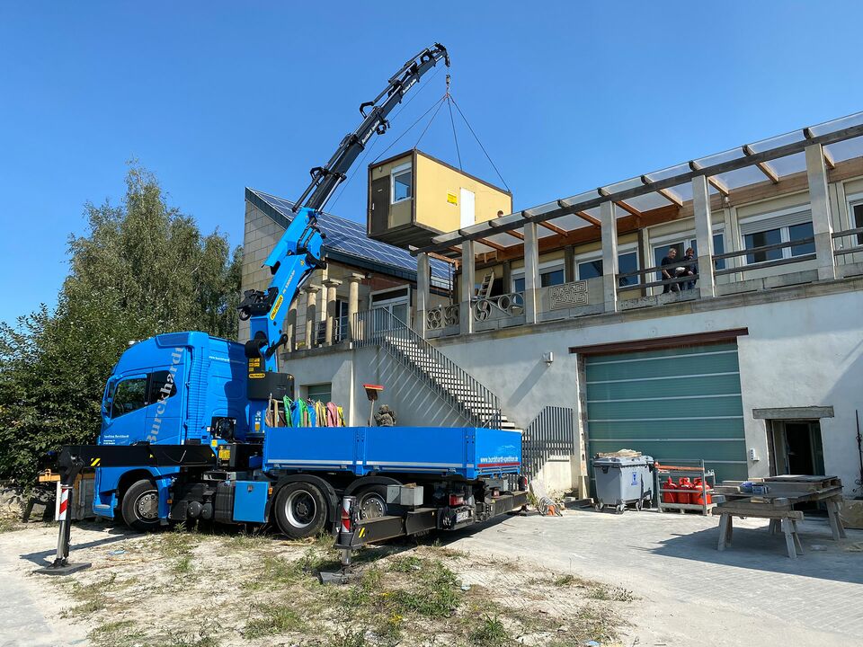 Ein blauer Kran hebt Baumaterialien auf das Dach eines Gewerbegebäudes bei strahlendem Sonnenschein.