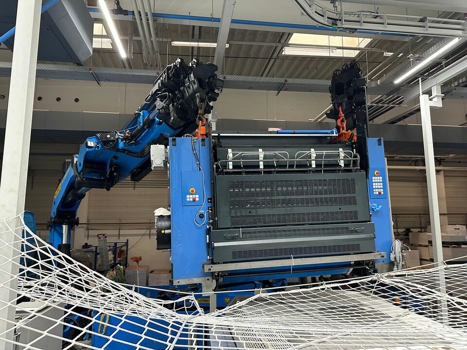 Ein blauer Kran hebt eine schwere Industriemaschine in einer Produktionshalle, umgeben von Sicherheitsnetzen und moderner Beleuchtung.