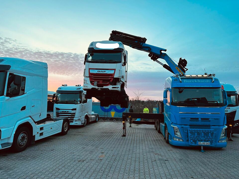 Ein blauer Kran hebt ein weiß-rotes LKW-Führerhaus zwischen geparkten LKWs, vor dem Hintergrund eines farbenfrohen Sonnenuntergangs, auf einen Transporter.