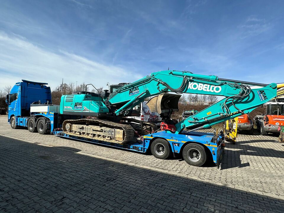 Transport eines Baggers mit Tieflader Ein blauer Tieflader-LKW transportiert einen großen, türkisfarbenen Bagger der Marke Kobelco auf einem Industriehof bei sonnigem Wetter.