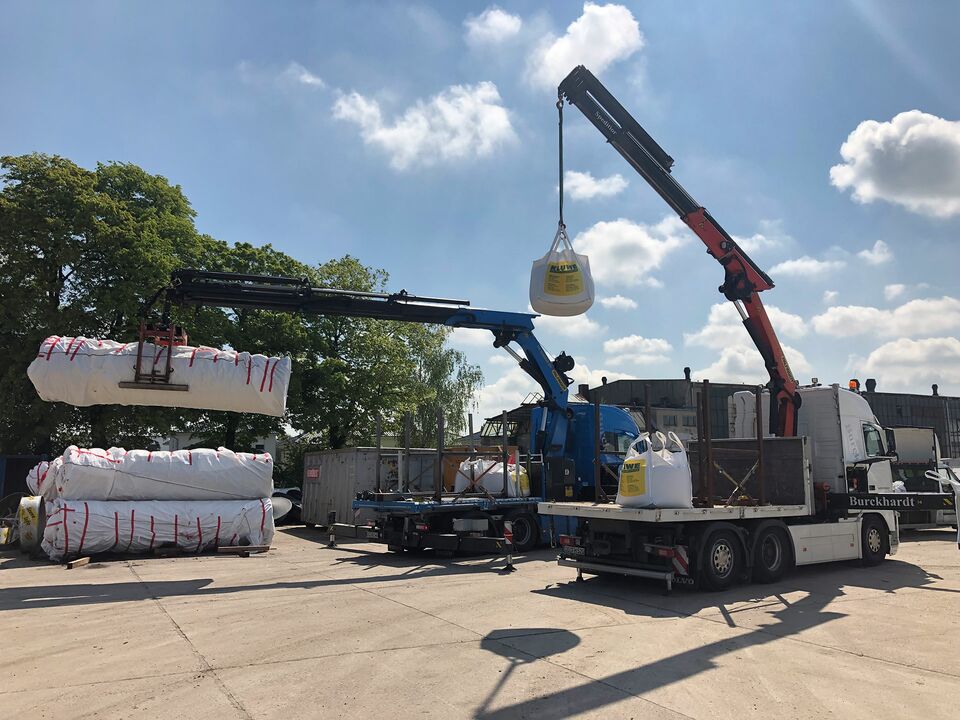 Zwei Kräne auf LKWs heben Baumaterialien wie Big Bags und Isolationspakete auf einem Lagerplatz an, bei klarem Himmel und Sonnenschein.