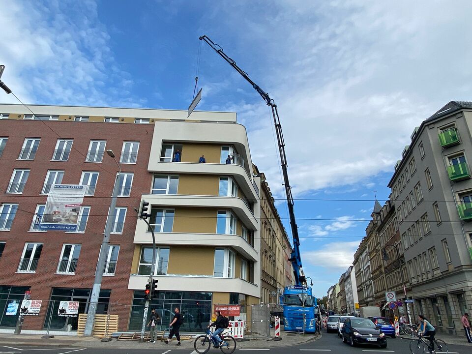 Ein blauer Kran hebt Bauteile auf das Dach eines modernen Wohngebäudes an einer belebten Straßenecke, umgeben von Fußgängern und Radfahrern.