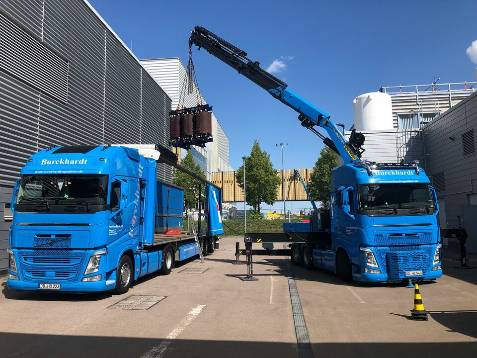 Zwei blaue LKWs mit Kränen heben eine schwere Last auf einem Industriehof an, umgeben von modernen Gebäuden und bei strahlendem Sonnenschein.