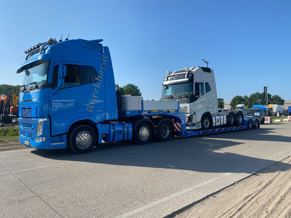 Ein blauer LKW transportiert ein weißes LKW-Führerhaus auf einem Tieflader über eine asphaltierte Straße bei strahlendem Sonnenschein.
