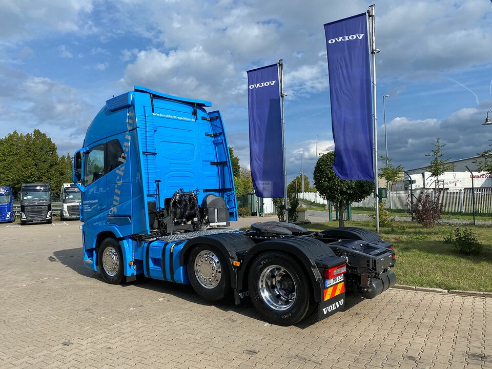 Ein blauer Volvo-LKW steht auf einem gepflasterten Platz, flankiert von Volvo-Flaggen und umgeben von Bäumen und weiteren LKWs.