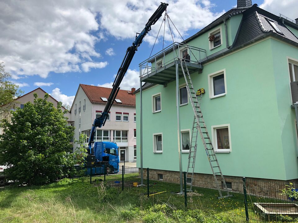 Ein blauer Kran hebt Baumaterialien auf das Dach eines grünen Wohnhauses in einem Wohngebiet.