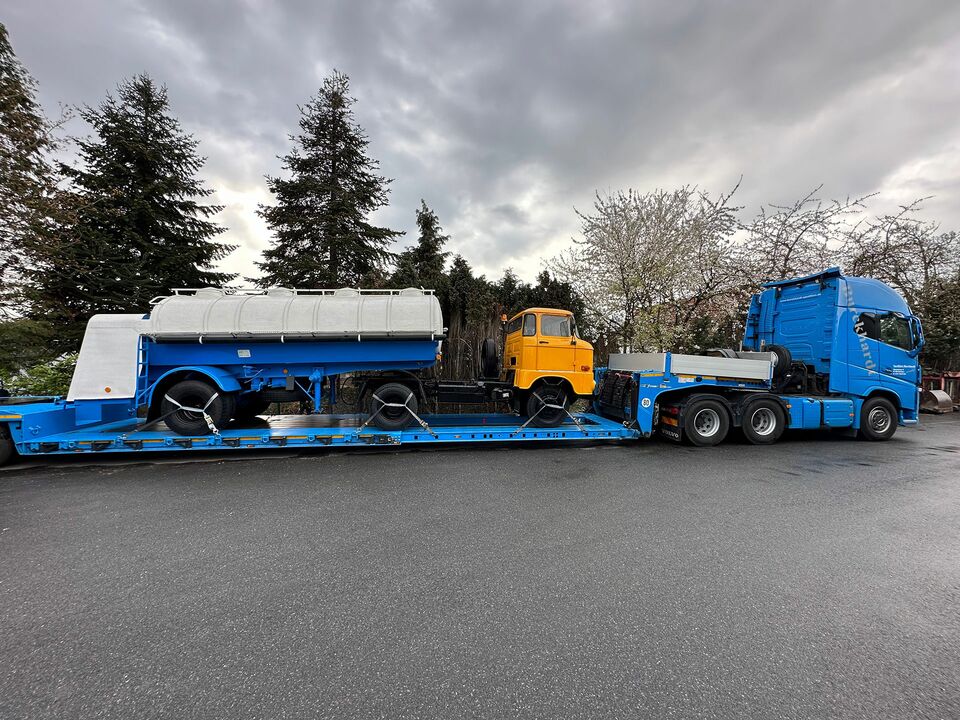Ein blauer Tieflader-LKW transportiert ein historisches gelbes Fahrzeug und einen Anhänger unter bewölktem Himmel, geparkt auf einer Straße mit Bäumen im Hintergrund.