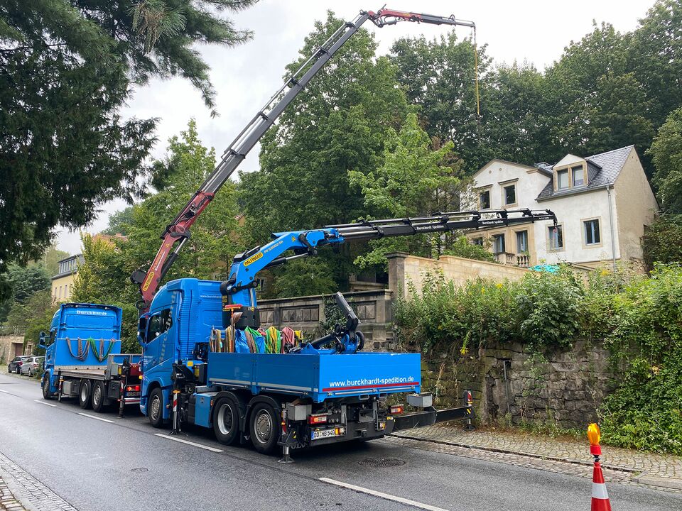 Zwei blaue LKWs mit Kränen heben Material in einer städtischen Wohngegend an einer engen Straße an, umgeben von Bäumen und Häusern.