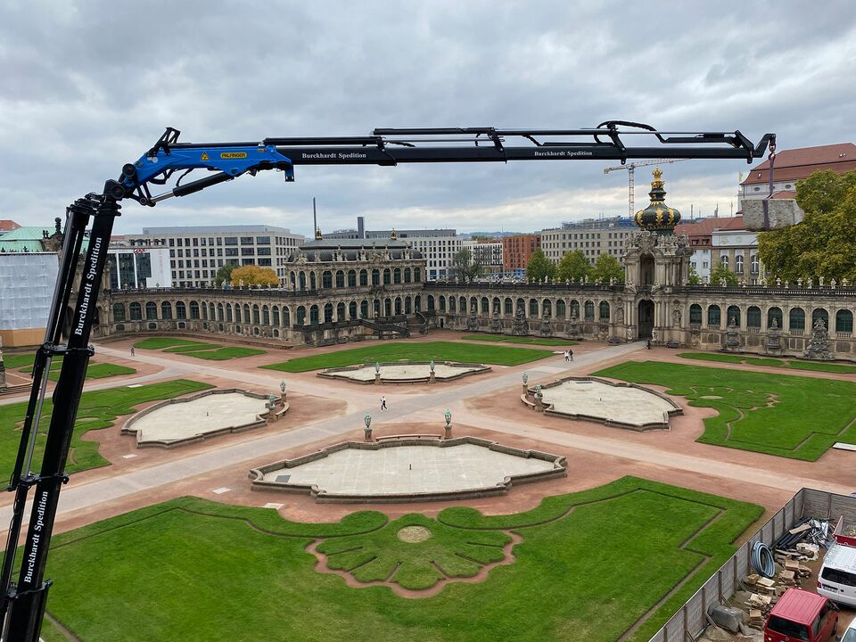 Ein blauer Kran arbeitet im historischen Zwinger-Park in Dresden, mit Blick auf die beeindruckende Barockarchitektur und gepflegte Gartenanlagen.