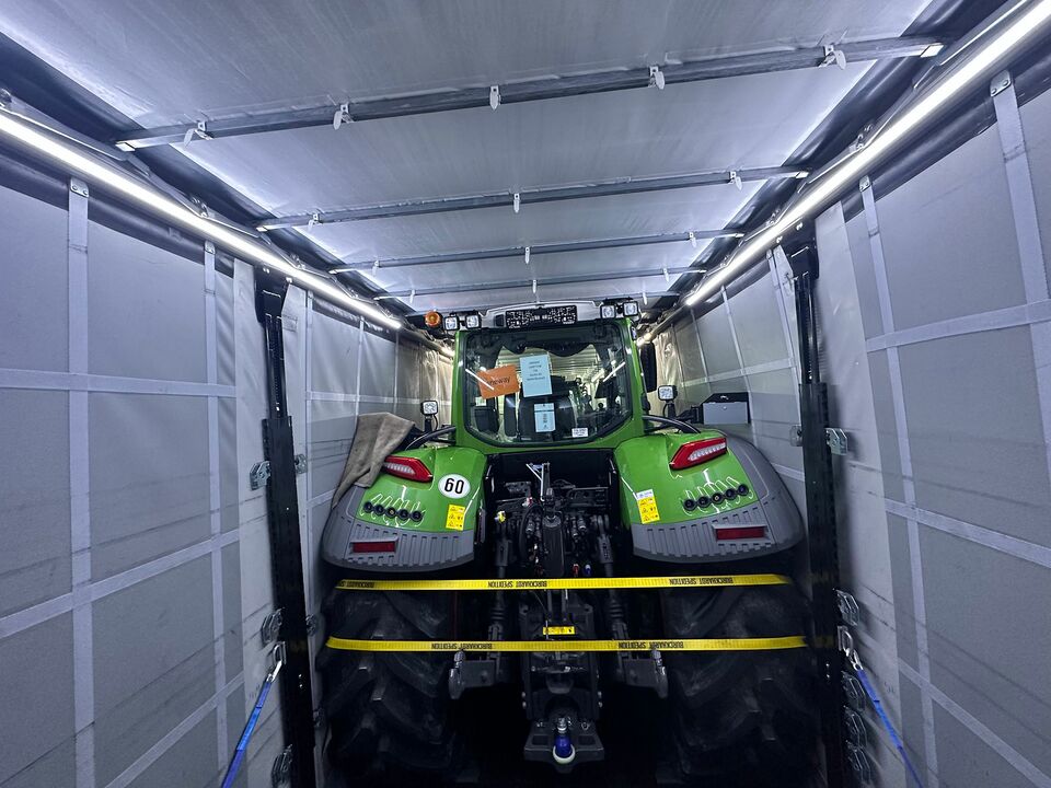 Ein grüner Traktor wird sicher mit Gurten in einem geschlossenen Anhänger transportiert, beleuchtet von LED-Lichtern.