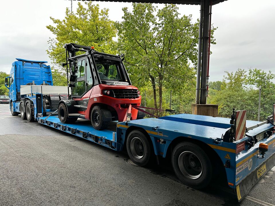 Ein roter Gabelstapler wird auf einem blauen Tieflader-LKW transportiert, geparkt unter einer Überdachung mit Blick auf Bäume im Hintergrund.